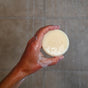 A hand holds a round bar of Badger Balm All-In-One Shampoo & Conditioner Bar - Cleanse & Hydrate under running shower water, with droplets visible on the bar and skin—ideal as a travel-friendly solid shampoo and conditioner for your daily routine.