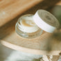 A jar of Little Seed Farm's Honey Lip Treatment with a white lid, slightly open to reveal the creamy balm inside, rests on a wooden surface in soft natural light. Blurred greenery appears in the foreground.