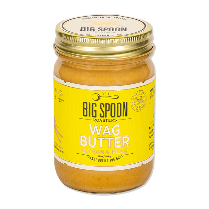 A glass jar of Big Spoon Roasters Banana Flax Wag Butter, a nutritious peanut butter for dogs, with a yellow label and gold lid.