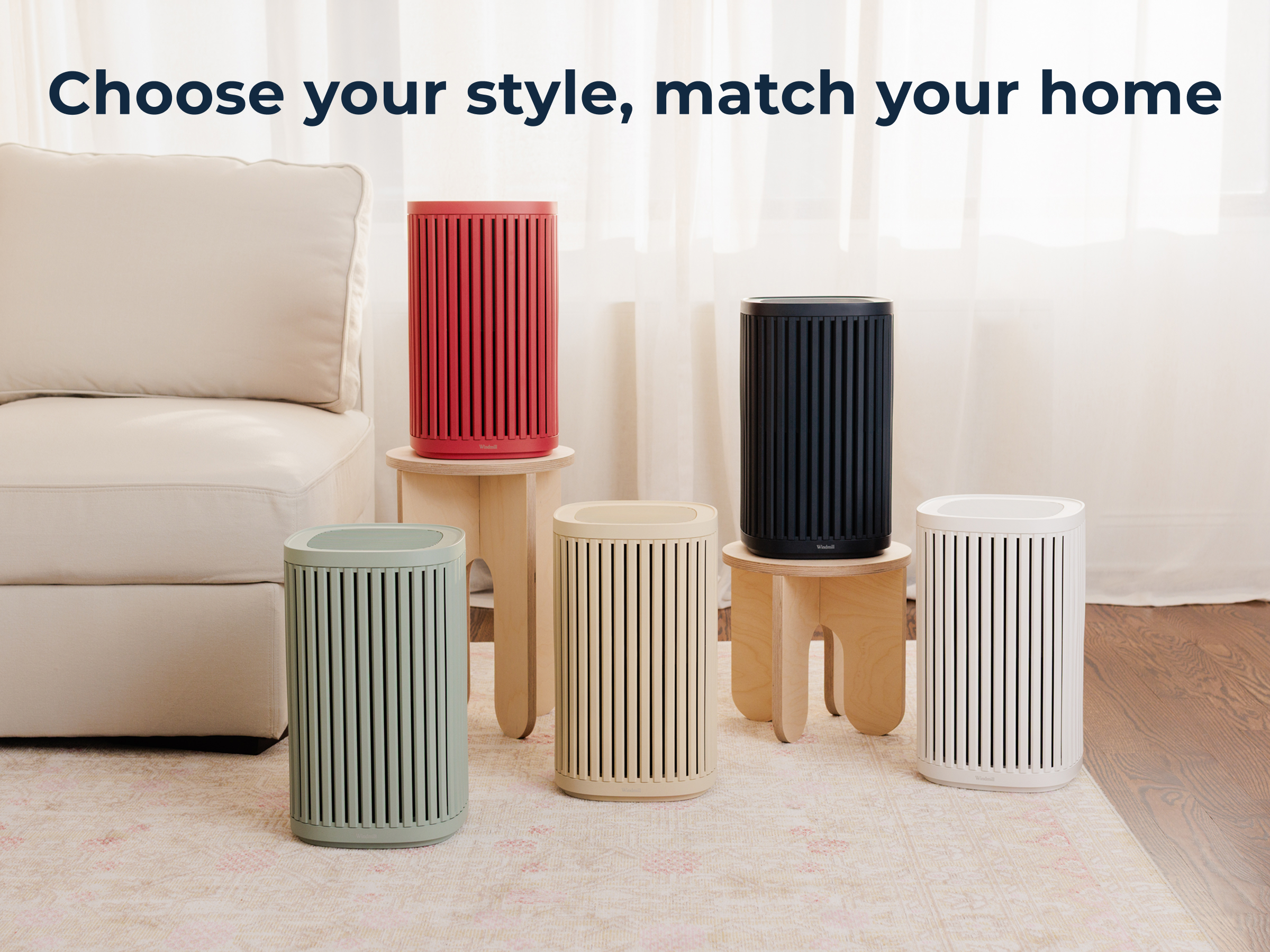 Five cylindrical Windmill Air Purifiers by Windmill Air, each in a different color, are displayed on stools and the floor in a living room, delivering advanced HEPA purification for your space. Text above reads: Choose your style, match your home.