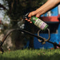 A person sprays Gear Hugger Heavy Duty Degreaser onto a metal garden tool outdoors, with green grass in the foreground and a blue and red building in the background.
