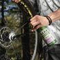 Outdoors among trees, a person sprays Gear Hugger Heavy Duty Degreaser onto a bicycle wheel, focusing on the disc brake, to clean it.