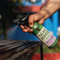 A hand sprays Gear Hugger Heavy Duty Degreaser onto a metal grill outdoors, with greenery and a blue building blurred in the background.