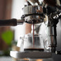 A Pure Over Espresso Cup Set holds rich, dark espresso as it pours from the machine, creamy crema forming on top. The softly blurred background highlights the artful coffee-making process and elegant glass cups.