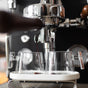 Close-up of the Pure Over Espresso Cup Set in use: an espresso machine pours rich, dark coffee with crema into clear glass cups from metal spouts.