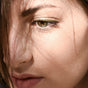 A woman's face in sunlight, her brown eye softly lined with Ere Perez Jojoba Eye Pencil. Shadows from her hair fall across her eye, nose, and cheek, highlighting her calm, thoughtful expression.