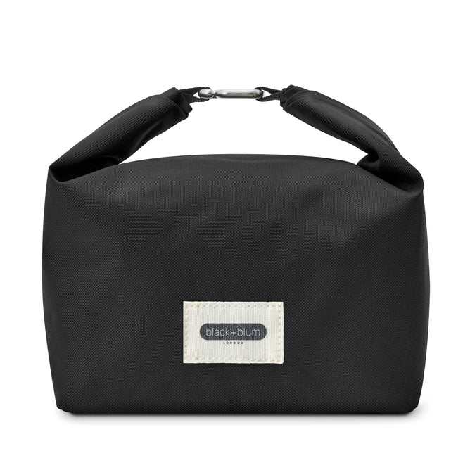 The Black + Blum Lunch Bag features black fabric, a light label with “black+blum London” on the front, and a rolled top secured by a metal clasp—ideal for carrying your lunch or everyday snacks.