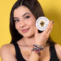 A woman with long dark hair holds a round, plastic-free NOPALERA Moisturizing Botanical Bar near her face, smiling slightly. She wears a black top and colorful woven bracelets against a yellow background.
