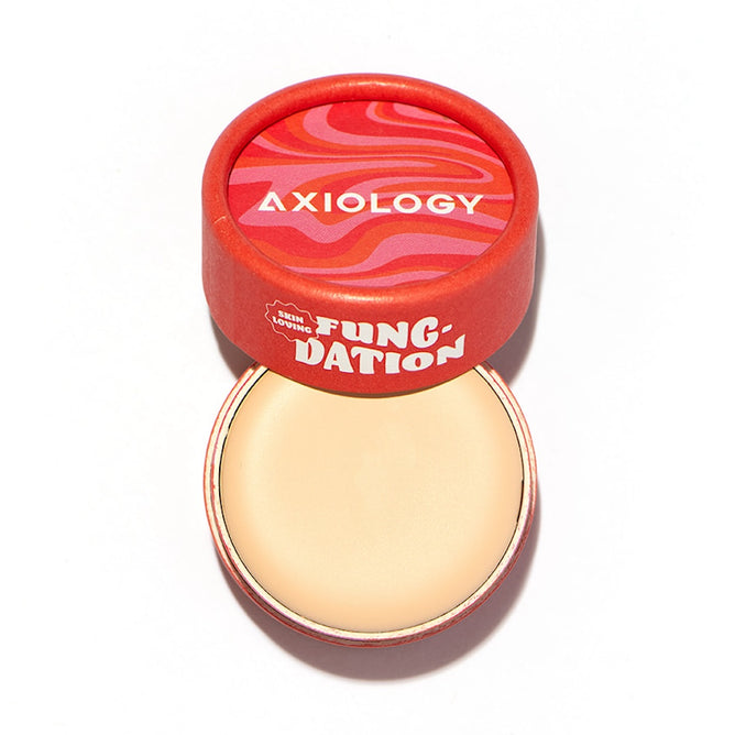 A round red container labeled Axiology with a swirled lid holds Fungdation – Mushroom Foundation, a pale cream hydrating base. The open container displays the smooth makeup inside.