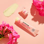 A tube and small jar of Sunniemade Nourishing Lip Balm - Blossom Butter, infused with moisturizing baobab oil, rest on a pink surface surrounded by pink flowers and a swipe of balm. The sustainable paper squeeze tube enhances the luxurious presentation of this lip balm from Sunniemade.