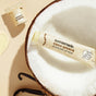 A tube of Sunniemade Hydrating Lip Balm - Coco Quench, enriched with nourishing cocoa butter and presented in recyclable packaging, is displayed inside a halved coconut, alongside a small coconut & vanilla jar and vanilla beans on a beige background.