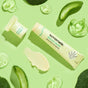 A tube of Sunniemade Healing Lip Balm - Hello Aloe, with its cap off, is displayed on a light green background and surrounded by aloe vera slices, gel, and leaves—beautifully showcased in recyclable packaging for an eco-friendly touch.