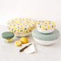 Esembly Baby’s Reusable Bowl Covers made from upcycled materials, 4-pack—two lemon print and two solid green—are shown on bowls near a glass jar, lemons, a knife, and napkin, all displayed on a white marble surface.