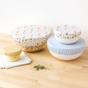 Four bowls topped with Esembly Baby’s Reusable Bowl Covers made from Upcycled Materials (4-Pack) sit on a wooden surface, with herbs and a white cloth nearby, showcasing stylish, eco-friendly food storage.