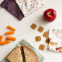 A sandwich in an Esembly Baby Reusable Snack & Sammy Food Pouch, an apple, carrot sticks, crackers spelling EAT, and a second pouch from the 2-pack are arranged on a white surface.