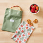 A sandwich in an Esembly Baby Reusable Snack & Sammy Food Pouch sits beside strawberries, three pretzels, and a strawberry-patterned cloth on a light wood surface for a waste-free lunchbox.