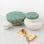Three bowls covered with Esembly Baby’s Reusable Bowl Covers made from upcycled materials (4-pack)—some in patterned green, one in plain white—rest on marble, with a small jar, wooden spoon, and yellow checkered napkin nearby.