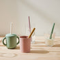 Four drinks with Esembly Baby’s Reusable Silicone Sipper Straws (4-pack, upcycled storage pouch) rest on wood: a green sippy cup, pink mug, clear water glass, and clear milk glass—each showcasing sustainable sipping.