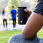A person holds an Elemental Stainless Steel Tumbler with Ceramic Lid, 12oz, while sitting on grass in a park as two people walk hand in hand in the blurred background.
