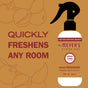 A Mrs. Meyers Room Freshener spray bottle is centered on a brown background with apple illustrations. Large text reads, Quickly freshens any room with this essential oil air freshener.