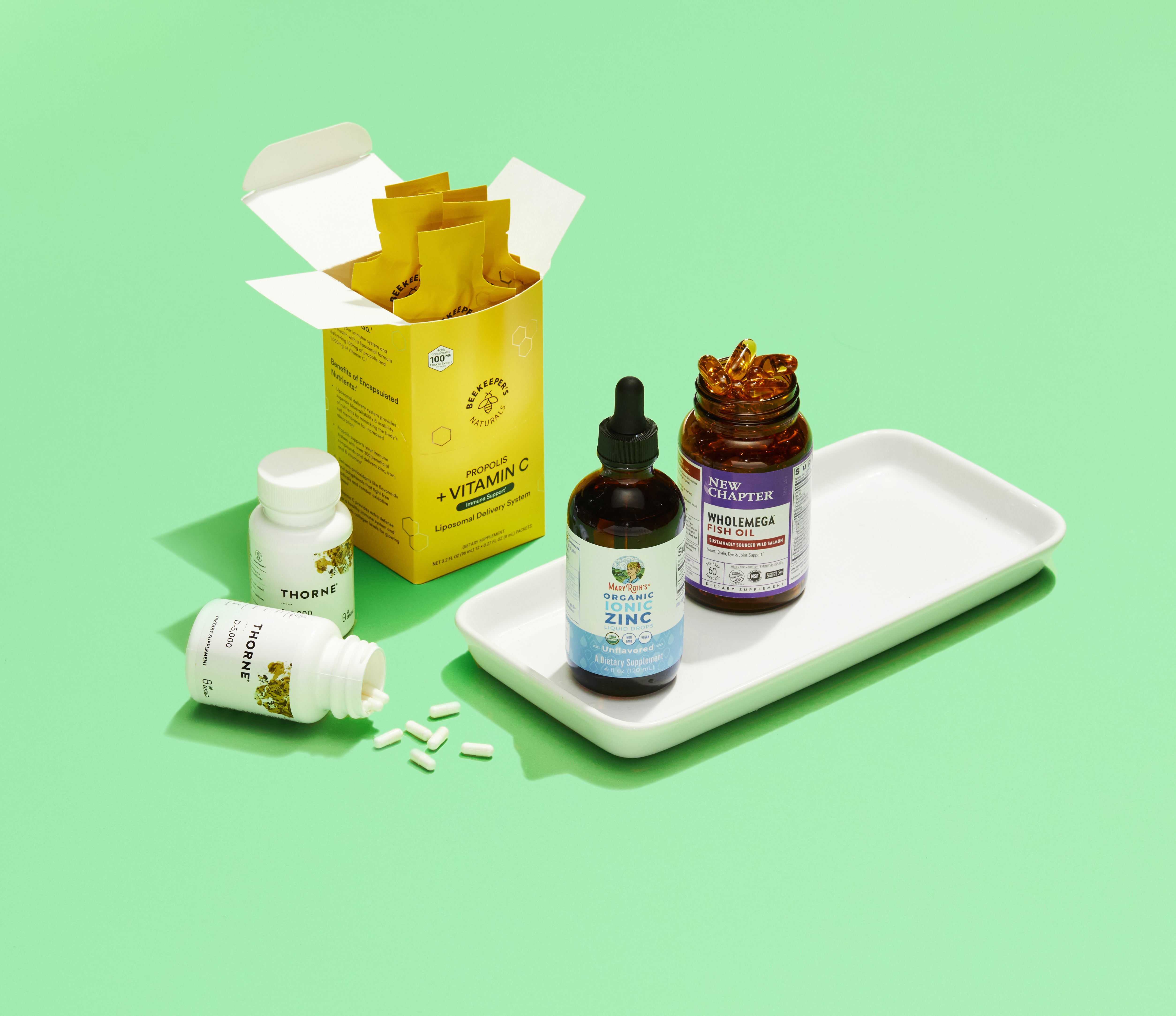 A box of vitamin C packets, two bottles of Thorne supplements with white capsules, a zinc liquid dropper bottle, and a bottle of softgel supplements are arranged on a white tray against a green background.