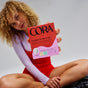A woman with curly blonde hair in a red and white outfit sits cross-legged, holding up a box of Cora Made to Move Pad - Sample, highlighting its You-Shaped fit for reliable leak protection. The Cora product is clearly visible and in focus.