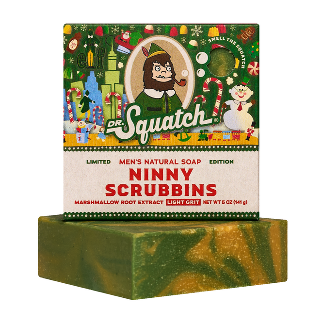 A green and yellow swirled Dr. Squatch Ninny Scrubbins Bar Soap, inspired by Buddy the Elf and made with marshmallow root extract, sits in front of its festive holiday box decorated with cheerful illustrations.