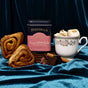 A tin of Spicewalla Deluxe Cinnamon Roll Hot Chocolate is shown with a cup topped with marshmallows, two cinnamon rolls, chocolate pieces, and cinnamon sticks, all arranged on rich blue velvet fabric.
