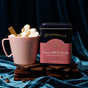 A pink mug of hot chocolate with marshmallows and a cinnamon stick sits on dark chocolate pieces next to a Spicewalla Deluxe Cinnamon Roll Hot Chocolate tin, with blue velvet fabric in the background.