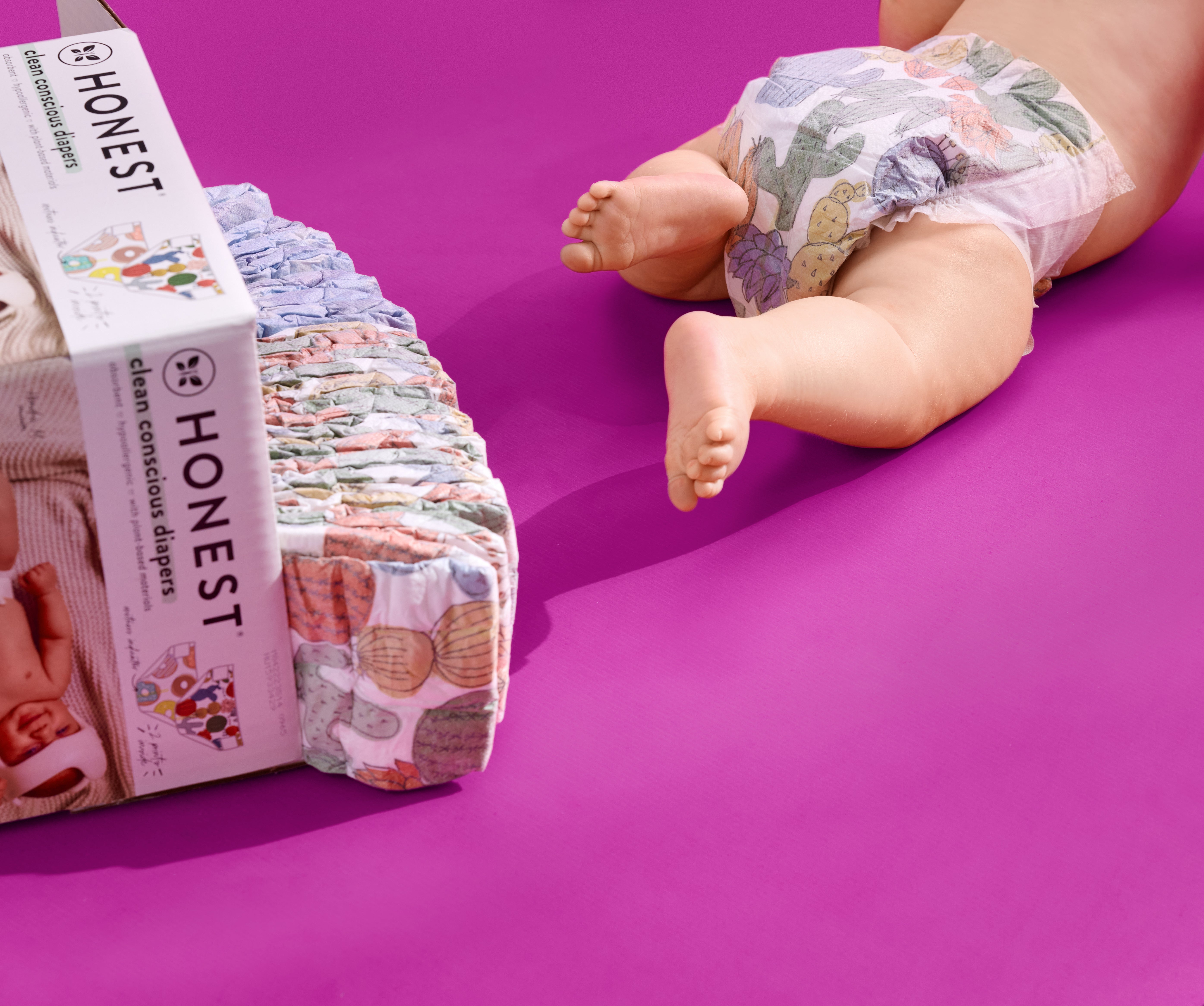 A baby wearing a colorful diaper lies on a bright pink surface next to a pack of Honest brand clean conscious diapers.