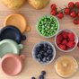 BEABA Glass Baby Food Jars with colorful lids store peas, blueberries, raspberries, and applesauce. Cherry tomatoes and pears sit nearby on a wooden surface; a few blueberries are scattered—ideal for homemade baby food storage.