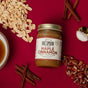 A jar of Big Spoon Roasters Maple Cinnamon Peanut & Pecan Butter sits on a red surface, surrounded by cinnamon sticks, peanuts, pecans, a bowl of syrup, and a spoonful of salt.