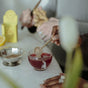 Someone stirs Wooden Spoon Herbs Lemon Ginger Immune Toddy sachet into a red drink in a glass, while another grates ginger nearby. A yellow immune support box and a metal dish sit on the table.