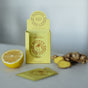 A yellow box labeled Wooden Spoon Herbs Lemon Ginger Immune Toddy - Sachets sits open on a table with elderberry, sliced lemon, ginger root and slices, plus a tea sachet—ideal for daily immune support.