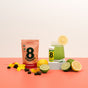 A glass of green drink with lemon, the 8Greens 10 Day Trial Kit – Gummies + Tablets, citrus slices, and a pink 8Greens pouch are neatly displayed on a red and white surface.