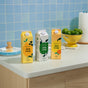 Three cartons of Grove Co. Dish Soap – Cuts Through Stuck-On Mess are on a kitchen counter beside limes, a paper towel roll, a dish brush, and plates, with blue tile behind them. Enjoy powerful grease cutting and natural fragrance.