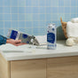 A person pours fabric softener from a Grove Co. carton into a Grove Co. Reusable Silicone Laundry Measuring Cup on a laundry room counter, with towels, a basket, and a wool dryer ball in the background.