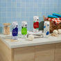 Four cartons of Grove Co. Laundry Detergent – 5-in-1 Enzyme Stain Fighting Power sit on a laundry room counter with towels, wool dryer balls, clothespins, and a wicker basket, set against a blue tiled wall.