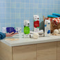 Four cartons of Grove Co. Laundry Detergent – 5-in-1 Enzyme Stain Fighting Power sit on a laundry room counter with towels, sponges, clothespins, and a basket; a hand reaches for one. A blue tiled wall is in the background.
