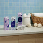 Three cartons of Grove Co. Delicate Care Laundry Detergent – Prevents Fading & Pilling are on a countertop with folded towels, wool dryer balls, a reusable silicone laundry measuring cup, and a wicker basket by a light blue tiled wall.