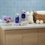 A hand pours Grove Co. Delicate Care Laundry Detergent – Prevents Fading & Pilling from a carton next to fabric softener, folded towels, dryer balls, and a basket. A Reusable Silicone Laundry Measuring Cup rests nearby on the tiled countertop.