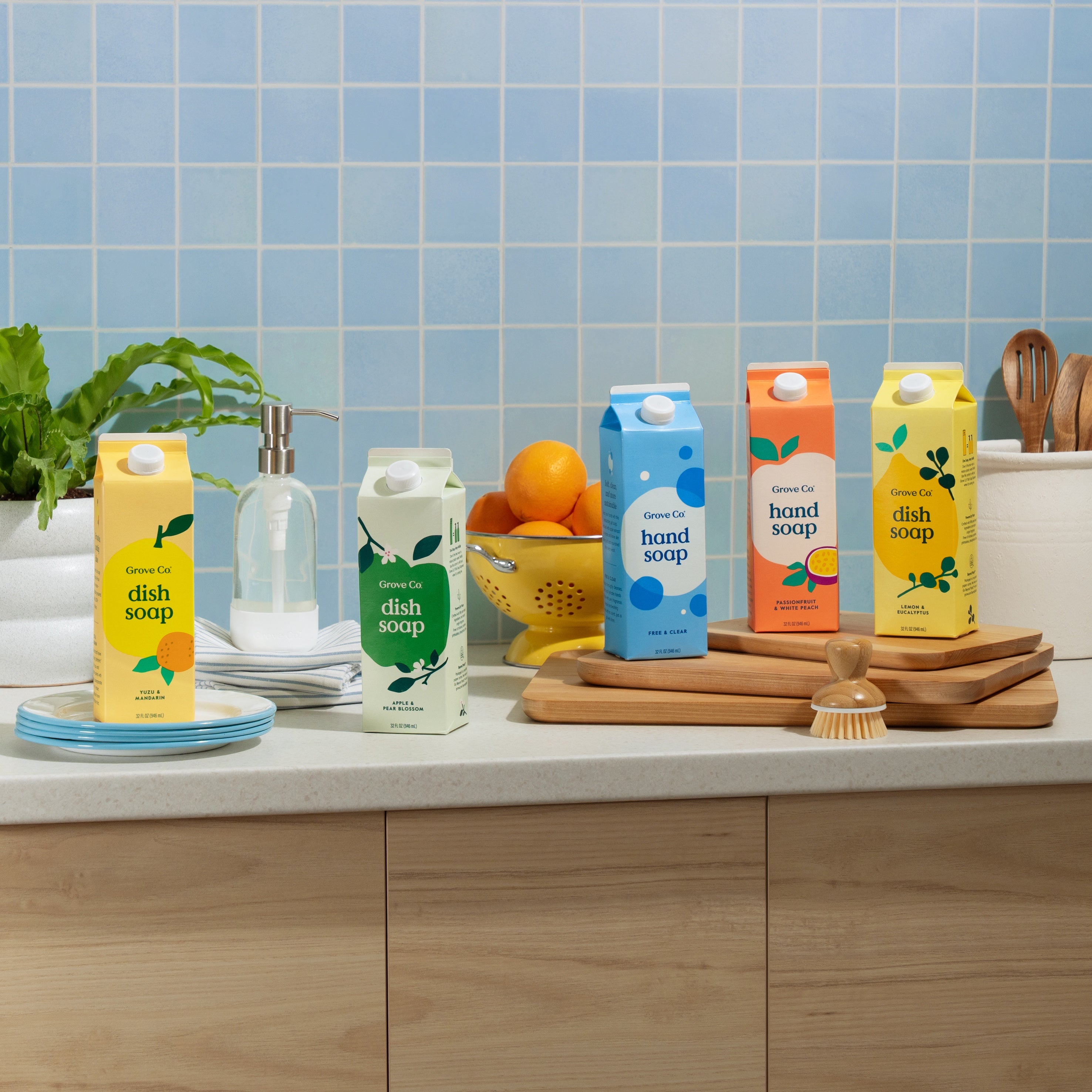 A kitchen counter displays Grove Co. Hand Soap – Hydrates & Moisturizes With Every Wash cartons in various colors, surrounded by dishes, a soap dispenser, a wooden brush, oranges in a bowl, a potted plant, and blue tiled wall backdrop.