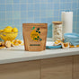 A bag of Grove Co. Dishwasher Detergent Packs – Triple-Action Clean, made with bio-based ingredients, sits on a kitchen counter next to a bowl of lemons, a scrub brush, stacked plates, blue bowls, a yellow towel, and a paper towel roll.