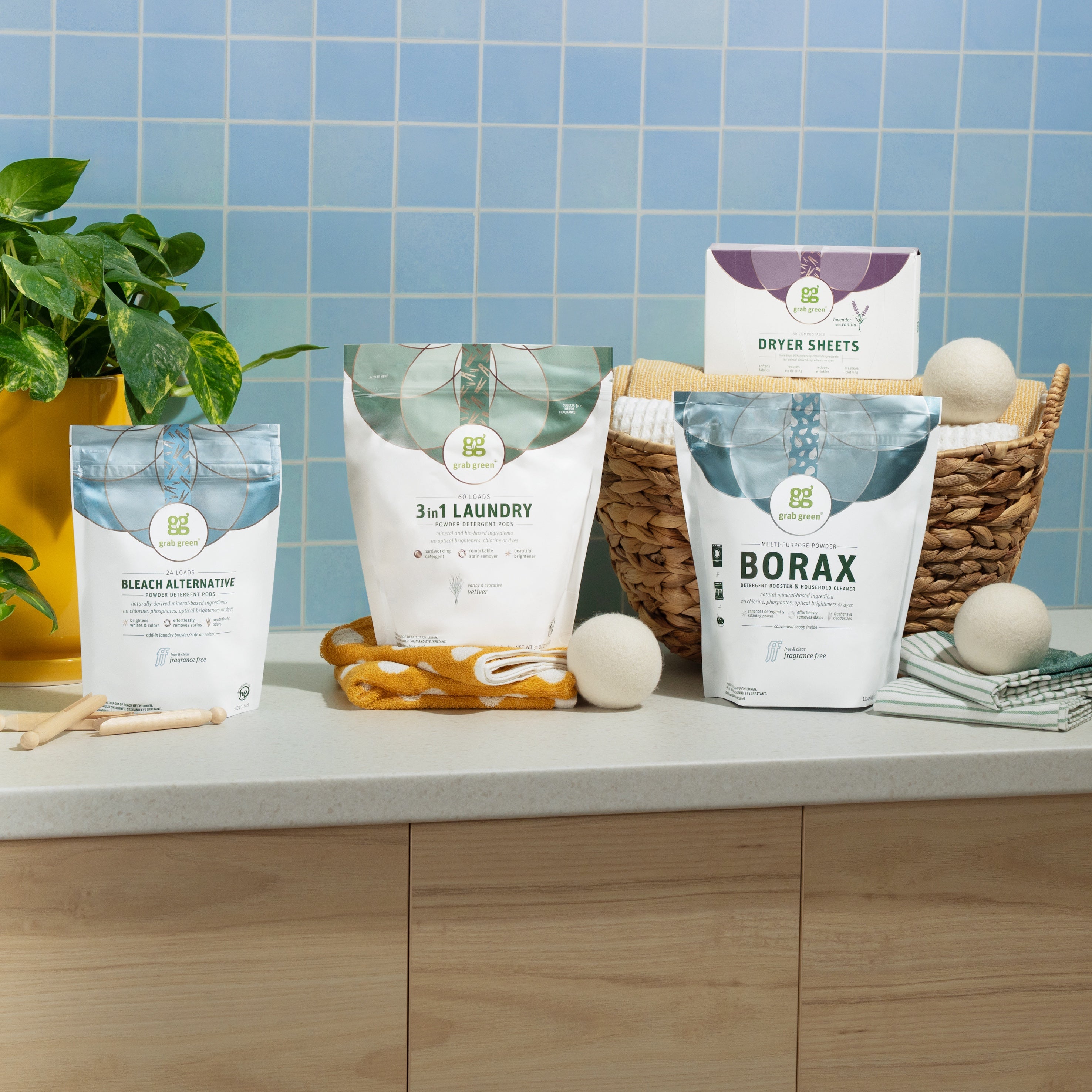 A countertop with grab green eco-friendly laundry products, including wool dryer balls, Bleach Alternative Pods, a small plant, and a wicker basket is set before a blue tiled wall. Packaging reads “3 in 1 Laundry” and “Dryer Sheets.”.