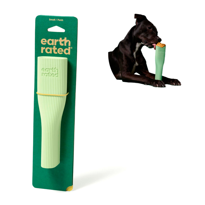 On the left is a packaged Earth Rated Enrichment Natural Rubber Chew Dog Toy. On the right, a black dog lies down, chewing this toy—filled with a tan treat—to promote behavioral development.