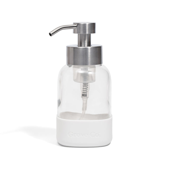 A Grove Co. Foaming Hand Soap Dispenser with a white silicone sleeve, featuring a clear glass body and stainless steel pump, designed with a refillable system to help reduce plastic waste, photographed on a plain white background.