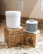 Two Epic Water Filters USA Pure XP Replacement Filter boxes sit on a granite counter; one contains a sealed white filter container, while the other reveals an exposed cylindrical Pure XP Replacement Filter cartridge with visible pleats—demonstrating the advanced water filtration technology behind these NSF certified filters.