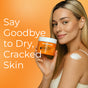A smiling woman applies Tree To Tub Moisturizing Coconut, Cocoa & Shea Butter Body Butter to her shoulder, holding the jar with “Say Goodbye to Dry, Cracked Skin” on an orange background.