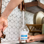 A person fills a reusable bottle with water and adds Meliora All-Purpose Home Cleaner Refill Tablets, with a dish rack and plates in the background.