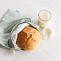 A round loaf with a crispy crust peeks from a checkered bread bag on white marble, alongside an open glass jar of creamy sourdough starter, completing the charming baking display of Esembly Babys Sourdough Proofing Kit.